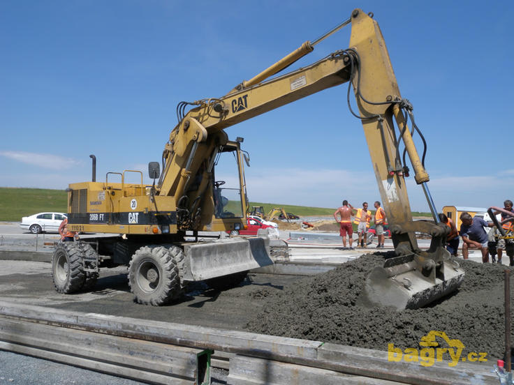 Kolové rypadlo Caterpillar 206B FT firmy Skanska DS rozhrnuje beton před finišerem